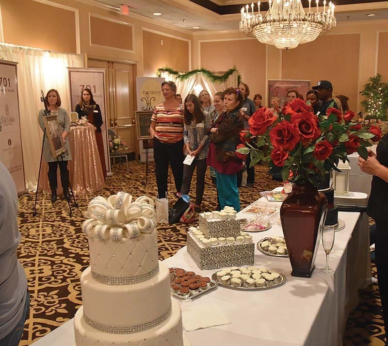 A tiered white wedding cake with a bow and rhinestone bands beside dessert tables in a ballroom, with guests listening to speakers.