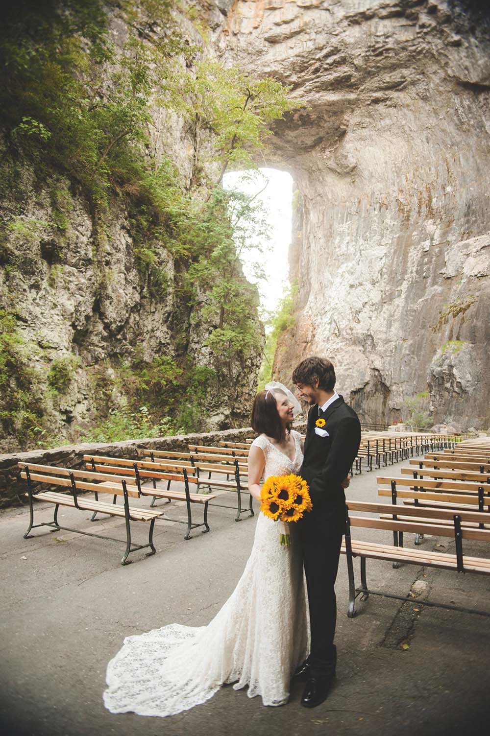 A bride in a lace gown with sunflower bouquet and a groom in a dark suit stand before a large natural rock arch and rows of wooden benches.