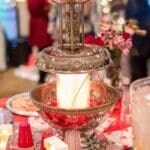 A tiered silver fountain with red liquid sits on a red tablecloth beside red cups, candy, and lit marquee letters.