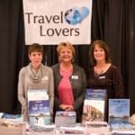 Three women stand behind a Travel Lovers booth with brochures for Africa, Central & South America, and island destinations.