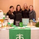Four women smile together behind an Arbonne booth with green tablecloth, "Enter to win" sign, and vegan products.