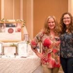 Two smiling women stand beside a display table with a framed "Tina Lane" sign and award plaques.