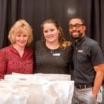 Three smiling people stand together behind a table of white gift bags against a black curtain.