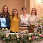 Four women stand smiling behind a table with floral garland, desserts, and a monitor showing a wedding photo.
