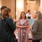 Three women chat in a warmly lit hotel ballroom foyer, with a man and a floral arrangement nearby.