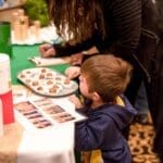 A young boy in a navy hoodie reaches toward a tray of bite-sized treats at a booth while an adult signs a clipboard.