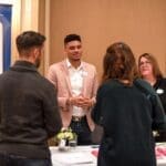 A man in a blush blazer and open collar talks with guests at a Hilton Garden Inn exhibitor table beside a banner.