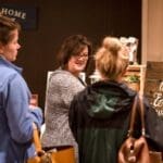 A smiling woman in glasses talks with two guests beside a rustic wooden sign reading "Love, laughter & happily ever after.