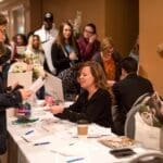 A woman seated at a white-draped registration table signs papers as attendees line up at a wedding show check-in.
