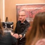 A bald chef in a black jacket and glasses holds up a skewer near a banner reading "we've always believed in putting our best foot forward.
