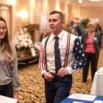 A man in an American flag-patterned shirt sleeve and suspenders talks with a woman at a bridal expo booth.