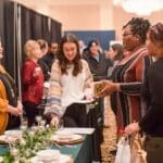 Guests chat with vendors at a bridal show booth decorated with greenery, glassware, and place settings in a hotel ballroom.