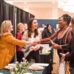 Two women shake hands at a bridal show booth, with a woman in a yellow blazer and guests nearby.