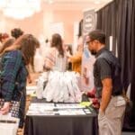 A man in glasses and a dark shirt looks at a vendor table with brochures and white gift bags at a bridal expo.