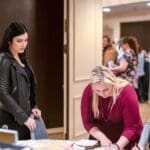 A woman in a black leather jacket stands at a table while a blonde woman in a maroon top works over it at an event.