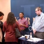 Three people talk at an event booth, with a navy banner reading "Wyndham" and "Mark" behind them.