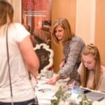 A woman in a patterned top helps guests at a registration table with a "Yes!" sign and eucalyptus centerpiece.
