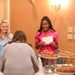 A woman in a pink polo holds a card beside a table of cookies while another woman in blue smiles nearby.