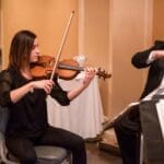 Two violinists in black formal attire perform side by side in a warmly lit room with music stands.