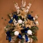 A white urn holds an arrangement of gold glitter poinsettias, navy velvet bows, white dahlias, delphinium, and ferns.