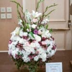 Large white and pink floral arrangement with lilies, roses, chrysanthemums and gladiolus in a pedestal vase, with a "Forest Flowers & Gift Gallery" placard.