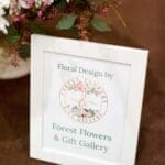 White framed sign reading "Floral Design by Forest Flowers & Gift Gallery" beside white flowers on a brown table.