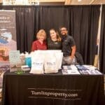 Three people stand smiling behind a Tomlin & Associates booth with a "Vacation Right At Home" banner and Tomlinproperty.com tablecloth.
