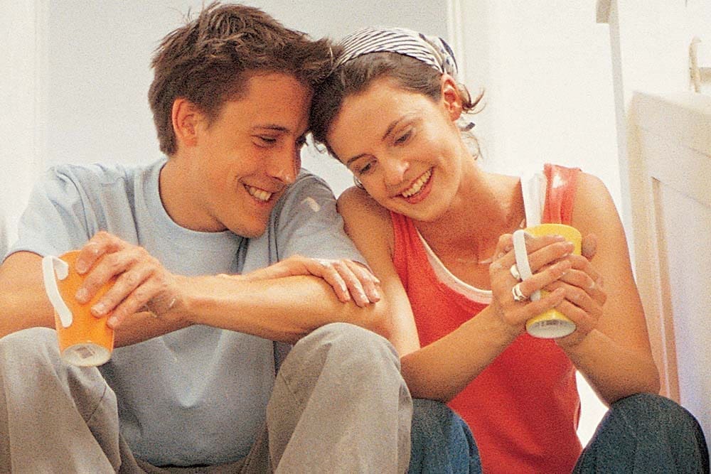 A smiling man in a light blue shirt and a woman in a red tank top sit close together, each holding a drink can.