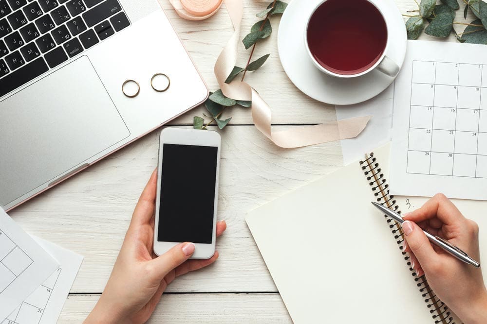Overhead view of hands holding a phone and writing in a notebook beside a laptop, rings, tea, and a calendar.