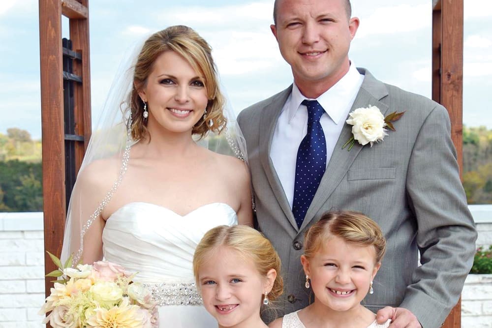 A bride in a strapless gown and veil and a groom in a gray suit pose with two smiling young girls outdoors.
