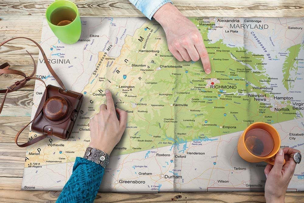 Overhead view of hands pointing at Richmond on a Virginia road map, beside two mugs and a vintage camera.