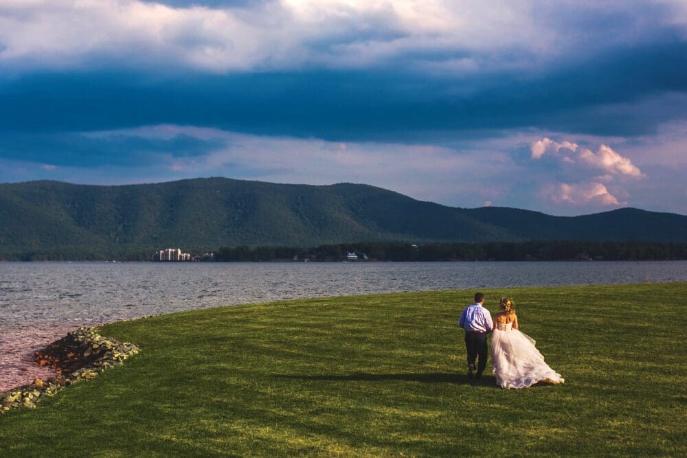 A couple in wedding attire walks hand in hand across a grassy lawn toward a lake and forested mountains under dramatic clouds.