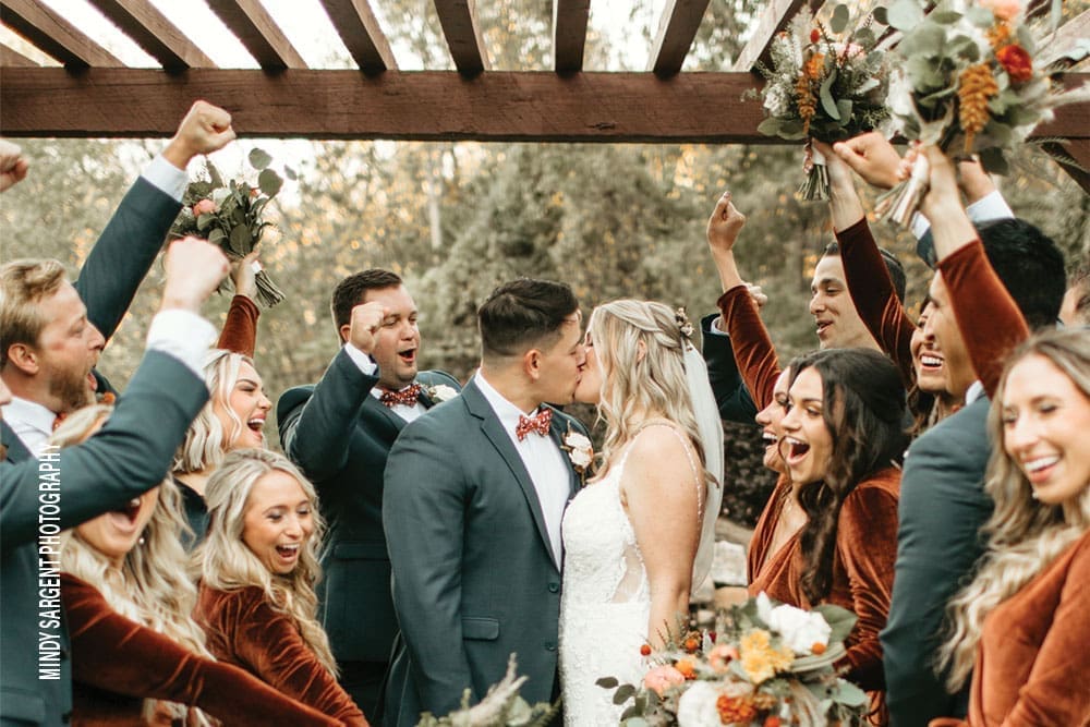 A bride and groom kiss under a wooden pergola while cheering bridesmaids and groomsmen raise bouquets and fists around them.