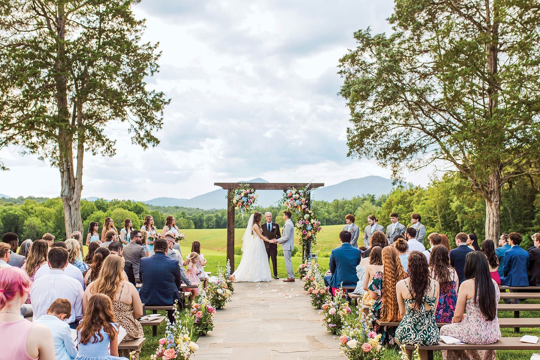 An outdoor ceremony with a couple exchanging vows under a wooden arch, guests seated on benches along a stone aisle, mountains behind.