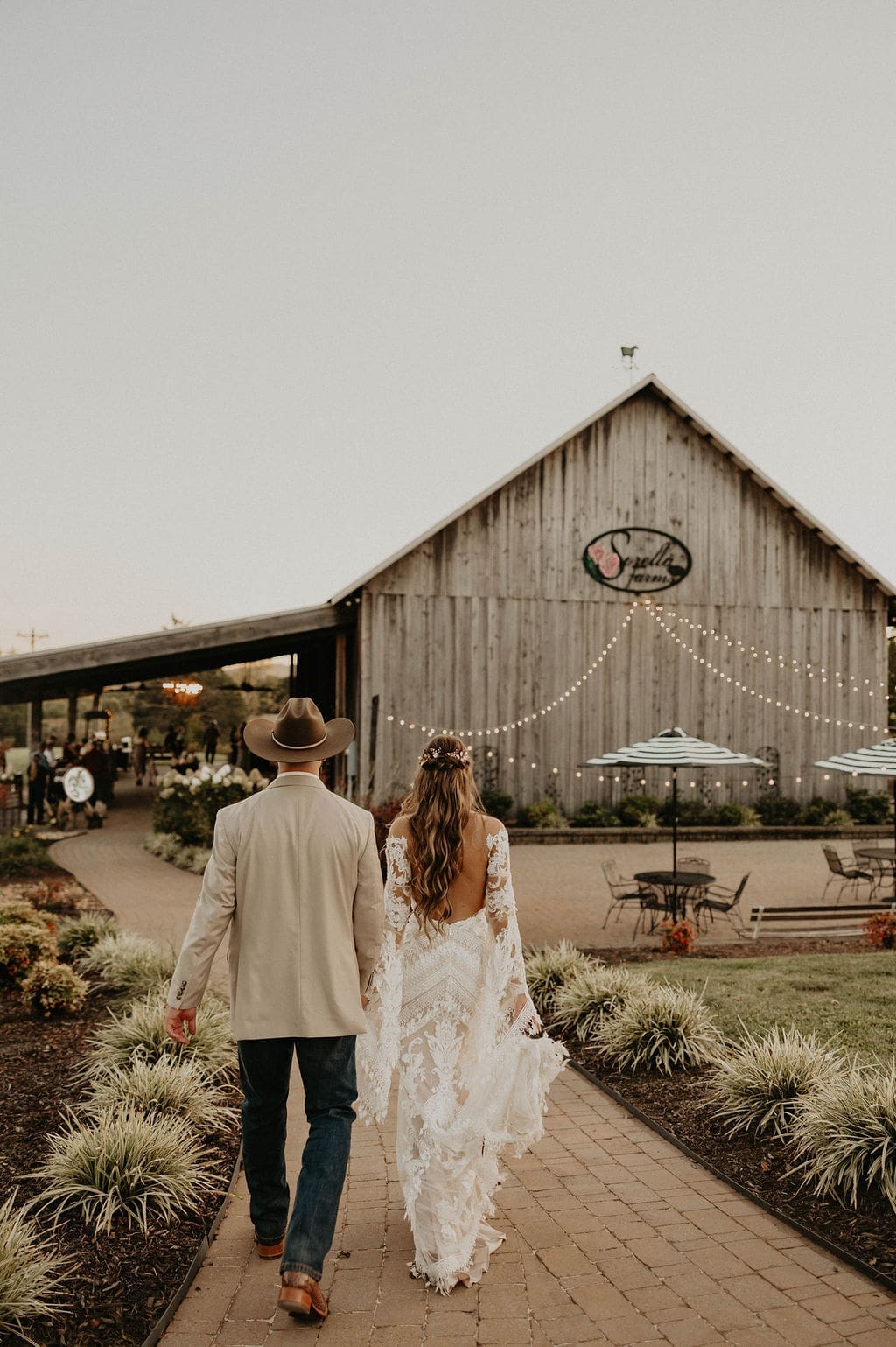 A couple in a cowboy hat and tan jacket and a lace-backed gown walks down a brick path toward a weathered barn with string lights.