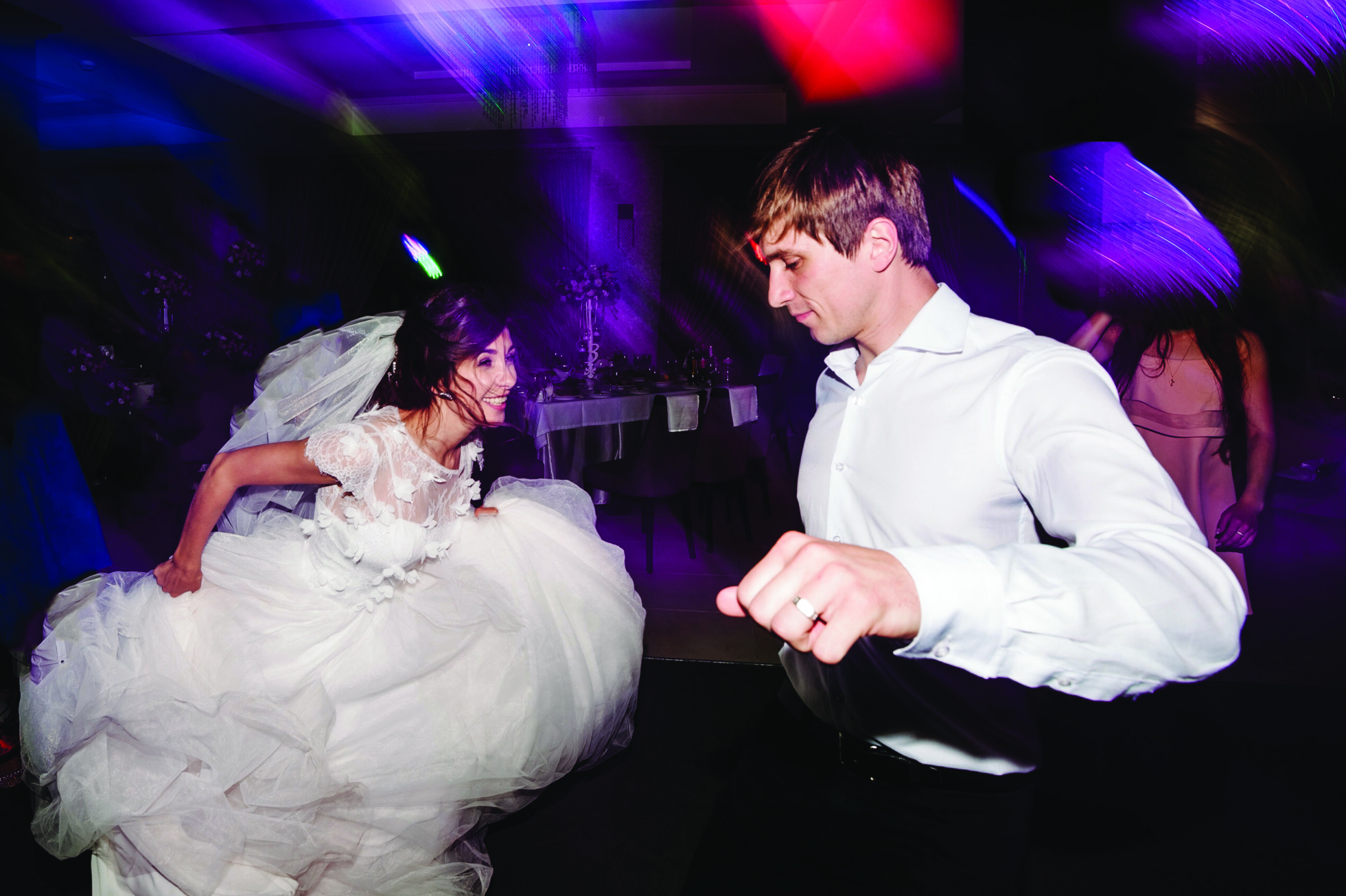 A bride in a tulle gown and veil dances energetically beside a groom in a white shirt under purple reception lighting.